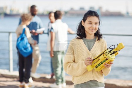 Photo for Waist up portrait of Asian teenage girl holding robot model and smiling at camera during outdoor engineering class with kids in background, copy space - Royalty Free Image