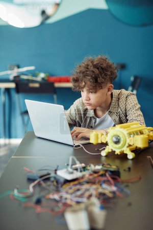 Téléchargez les photos : Portrait vertical d'un jeune garçon aux cheveux bouclés utilisant un ordinateur portable à l'école pendant un cours d'ingénierie et un robot de programmation - en image libre de droit