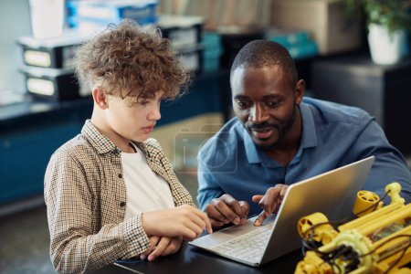 Téléchargez les photos : Portrait d'un enseignant noir aidant un jeune garçon à construire un robot pendant un cours d'ingénierie à l'école et utilisant un ordinateur portable pour programmer ensemble - en image libre de droit