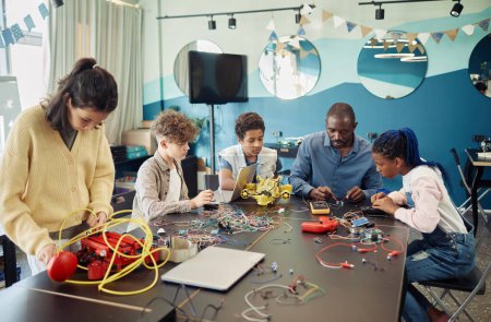 Téléchargez les photos : Groupe diversifié d'enfants construisant des robots avec l'aide d'un enseignant masculin pendant les cours d'ingénierie à l'école - en image libre de droit
