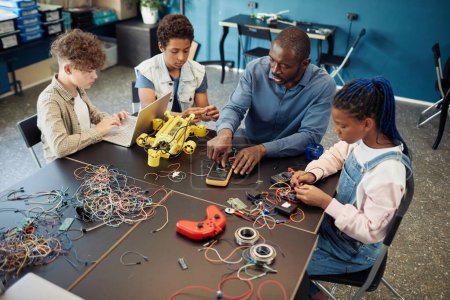 Téléchargez les photos : Vue grand angle à divers groupes d'enfants construisant des robots pendant les cours d'ingénierie à l'école avec l'aide d'un enseignant masculin - en image libre de droit