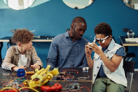 Téléchargez les photos : Portrait de jeune garçon noir construisant des robots en classe d'ingénierie avec l'aide d'un enseignant masculin - en image libre de droit