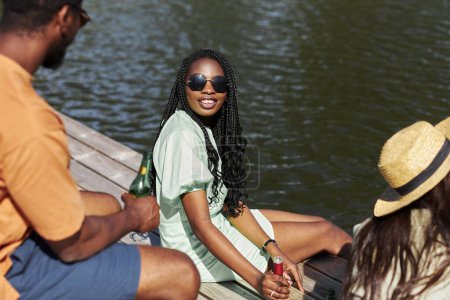 Téléchargez les photos : Portrait de jeune femme noire portant des lunettes de soleil et bavardant avec des amis à l'extérieur tout en se relaxant avec des boissons à l'eau - en image libre de droit