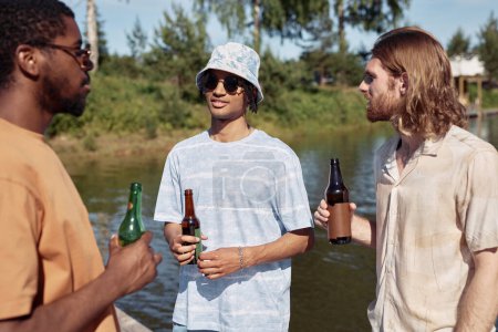 Foto de Cintura hacia arriba retrato de tres hombres jóvenes bebiendo cerveza al aire libre y charlando en el entorno de verano - Imagen libre de derechos