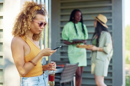 Photo for Side view portrait of young woman holding smartphone during barbeque party outdoors in Summer, copy space - Royalty Free Image