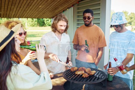 Foto de Diverso grupo de amigos asando carne durante la fiesta de barbacoa al aire libre en verano y bebiendo cerveza - Imagen libre de derechos