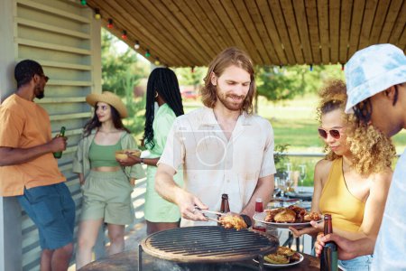 Téléchargez les photos : Portrait taille haute d'un homme souriant grillant de la viande à l'extérieur pendant la fête barbecue d'été avec des amis - en image libre de droit