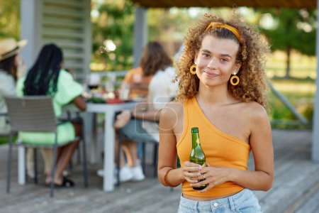 Foto de Retrato de la cintura hacia arriba de la joven rizada sonriendo a la cámara y sosteniendo la bebida durante la fiesta al aire libre con amigos en verano, espacio para copiar - Imagen libre de derechos