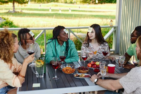 Téléchargez les photos : Groupe diversifié d'amis jouant Devinez qui joue assis à la table à l'extérieur en été - en image libre de droit