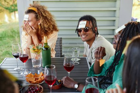 Téléchargez les photos : Groupe de jeunes gens riant et jouant Devinez qui joue assis à la table à l'extérieur en été - en image libre de droit