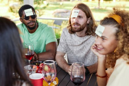 Téléchargez les photos : Jeune homme avec autocollant note sur la tête jouer Devinez qui jeu avec divers groupes d'amis à la fête en plein air - en image libre de droit