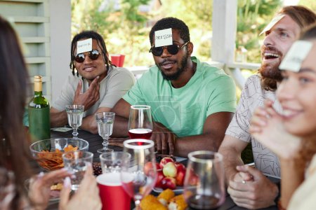Téléchargez les photos : Portrait d'homme noir avec autocollant note sur la tête jouer Devinez qui jeu avec divers groupes d'amis à la fête en plein air - en image libre de droit
