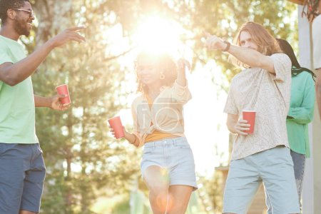 Foto de Diverso grupo de jóvenes bailando al aire libre bajo la luz del sol durante la fiesta de verano, destello de lente - Imagen libre de derechos