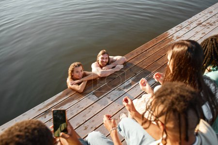 Foto de Vista de ángulo alto de pareja joven nadando en el lago con un grupo de amigos viendo desde el muelle y filmando video para redes sociales - Imagen libre de derechos