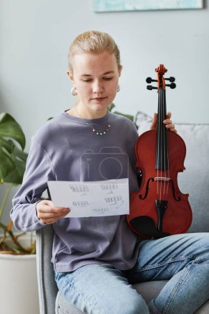 Photo for Vertical portrait of blonde young woman playing violin at home and holding music sheet - Royalty Free Image