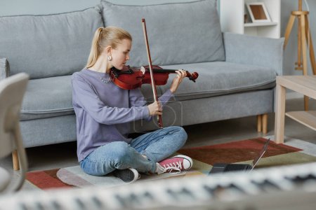 Téléchargez les photos : Portrait complet de jeune femme blonde jouant du violon à la maison tout en étant assis sur le sol dans un intérieur confortable, espace de copie - en image libre de droit
