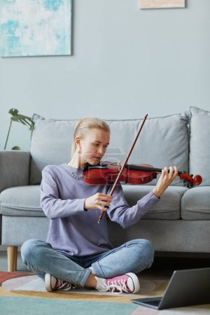 Téléchargez les photos : Portrait vertical complet de jeune femme blonde jouant du violon à la maison tout en étant assise sur le sol et en regardant une leçon de musique en ligne - en image libre de droit
