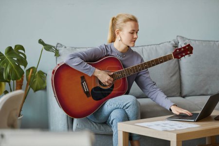 Foto de Retrato mínimo de una joven rubia tocando la guitarra en casa y componiendo música mientras está sentada en el sofá, espacio para copiar - Imagen libre de derechos