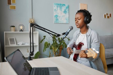 Foto de Retrato mínimo de una joven negra cantando al micrófono y tocando la guitarra en el estudio de grabación en casa, espacio para copiar - Imagen libre de derechos