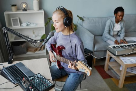 Téléchargez les photos : Portrait en grand angle d'une jeune femme blonde jouant de la guitare électrique et chantant au microphone dans un studio d'enregistrement à domicile, espace de copie - en image libre de droit