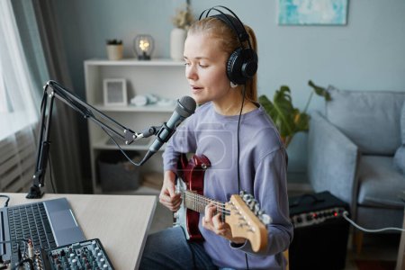 Téléchargez les photos : Portrait latéral de jeune femme blonde jouant de la guitare électrique et chantant au microphone dans un studio d'enregistrement à domicile, espace de copie - en image libre de droit