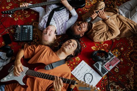 Foto de Vista superior de la banda de música sonriente tumbado en la alfombra adornada en el estudio mucis y mirando a la cámara - Imagen libre de derechos