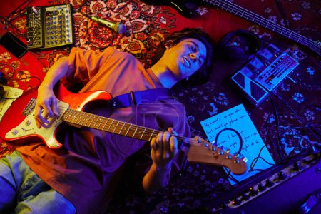 Photo for Top view shot of young musician playing guitar while lying on carpet lit by neon light - Royalty Free Image