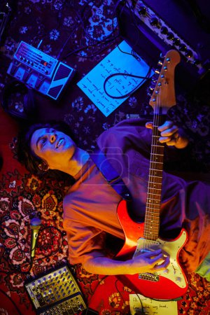 Photo for Top view shot of smiling young man playing guitar while lying on carpet in neon light - Royalty Free Image