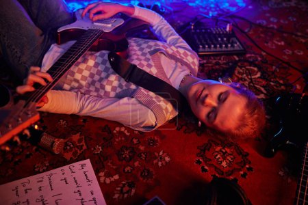 Téléchargez les photos : Gros plan d'une jeune femme jouant de la guitare allongée sur un tapis éclairé par des lumières rouges tamisées - en image libre de droit
