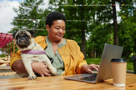 Téléchargez les photos : Portrait de femme noire senior utilisant un ordinateur portable dans un café en plein air avec chien carlin mignon - en image libre de droit