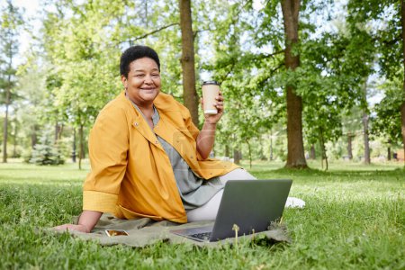 Téléchargez les photos : Portrait pleine longueur d'une femme mûre heureuse utilisant un ordinateur portable à l'extérieur tout en appréciant le pique-nique avec du café sur herbe verte, espace de copie - en image libre de droit