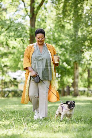 Vertical full length portrait of smiling black woman walking cute dog in park and enjoying coffee