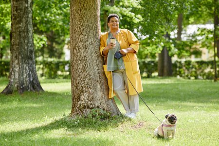 Téléchargez les photos : Portrait complet de la femme noire aînée appuyée sur l'arbre dans le parc tout en marchant chien mignon et en buvant du café, espace de copie - en image libre de droit