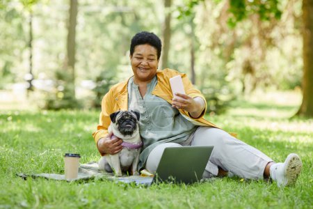 Foto de Retrato de cuerpo entero de una mujer negra madura tomando foto selfie con un perro mientras disfrutan de un picnic en el parque juntos, espacio para copiar - Imagen libre de derechos