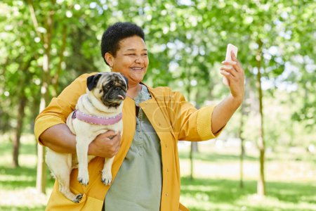 Téléchargez les photos : Portrait taille haute de femme noire mature prenant selfie photo avec chien dans le parc et souriant joyeusement - en image libre de droit