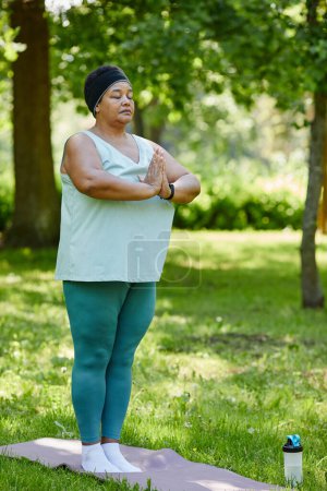 Téléchargez les photos : Portrait vertical pleine longueur de femme noire mature appréciant le yoga en plein air dans un parc vert et debout les yeux fermés faisant des exercices de respiration - en image libre de droit