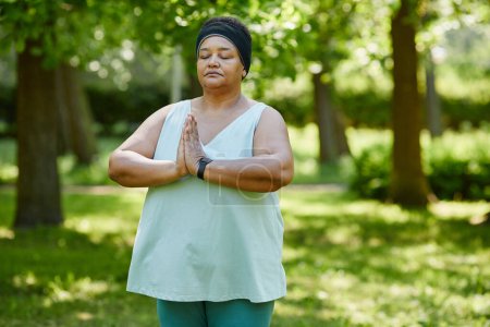 Téléchargez les photos : Portrait taille haute de femme noire mature faisant du yoga à l'extérieur dans un parc vert et tenant les mains ensemble en position de prière, copiez l'espace - en image libre de droit