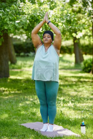 Téléchargez les photos : Portrait vertical pleine longueur de femme noire en surpoids faisant du yoga à l'extérieur dans un parc vert debout en position d'arbre - en image libre de droit