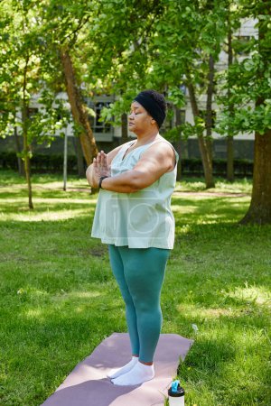 Téléchargez les photos : Portrait vertical pleine longueur de femme noire en surpoids faisant du yoga à l'extérieur dans un parc vert et méditant les yeux fermés - en image libre de droit