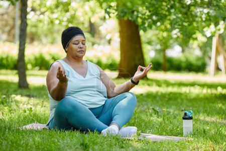 Foto de Retrato de cuerpo entero de una mujer negra con sobrepeso haciendo yoga al aire libre y meditando con los ojos cerrados sobre hierba verde - Imagen libre de derechos
