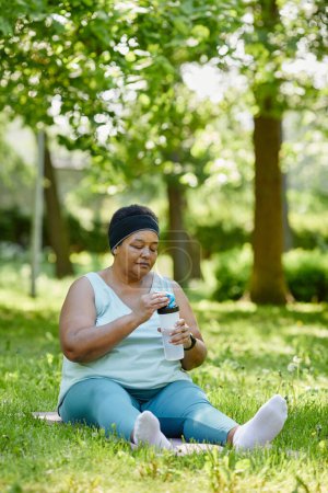 Foto de Retrato vertical de cuerpo entero de una mujer negra con sobrepeso haciendo ejercicio al aire libre y bebiendo agua para mantener la hidratación - Imagen libre de derechos