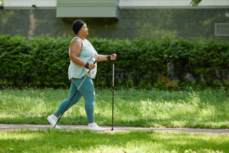 Photo for Side view full length of overweight black woman walking outdoors with nordic poles and enjoying cardio workout, copy space - Royalty Free Image