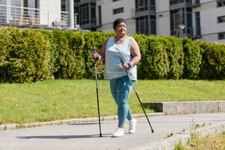 Téléchargez les photos : Portrait complet d'une femme souriante en surpoids marchant à l'extérieur avec des bâtons nordiques et profitant d'un entraînement cardio en plein soleil - en image libre de droit