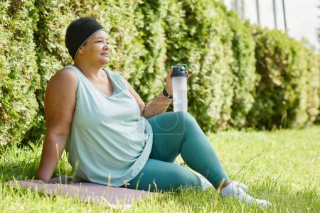 Téléchargez les photos : Pleine longueur vue latérale de femme noire en surpoids relaxant sur l'herbe tout en travaillant à l'extérieur avec une bouteille d'eau, espace de copie - en image libre de droit