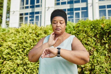 Téléchargez les photos : Portrait taille haute de femme noire mature regardant smartwatch et traçant pouls tout en prenant une pause dans l'entraînement en plein air - en image libre de droit