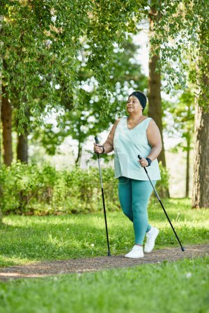 Téléchargez les photos : Portrait vertical complet de femme noire mature marchant avec des bâtons dans le parc pendant l'entraînement en plein air - en image libre de droit
