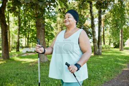 Téléchargez les photos : Portrait taille haute de souriante femme noire mature marchant avec des bâtons dans le parc pendant l'entraînement en plein air - en image libre de droit