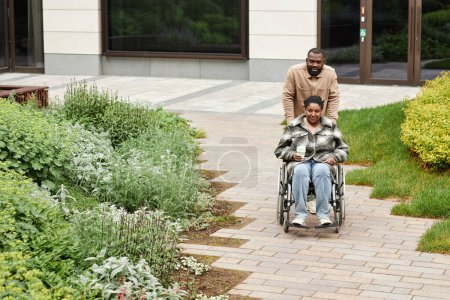 Téléchargez les photos : Portrait complet du couple noir moderne avec une jeune femme souriante en fauteuil roulant profitant d'une promenade dans le jardin de la ville, espace de copie - en image libre de droit