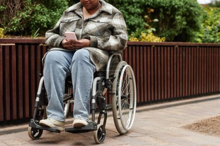 Photo for Cropped shot of black woman with disability using smartphone in city garden setting and wearing coat, copy space - Royalty Free Image