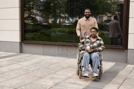 Téléchargez les photos : Minimal pleine longueur prise de vue de couple noir avec partenaire en fauteuil roulant regardant la caméra à l'extérieur dans le cadre de la ville, espace de copie - en image libre de droit